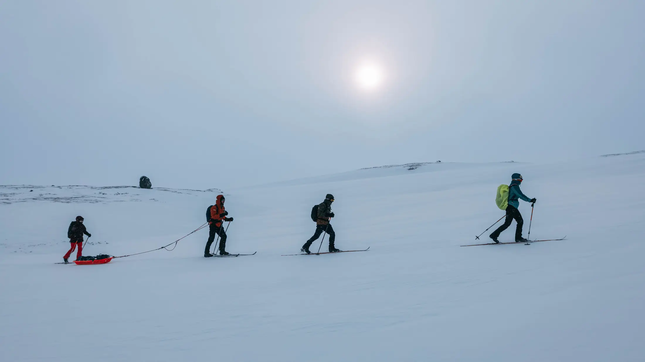 A line of skiers pulling their pulkas across a white plain under a hazy sun