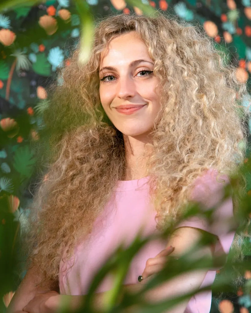 Portrait de Laura Lhuillier, longs cheveux bouclés, devant une fresque végétale